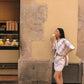 A woman stands on a sidewalk beside a textured beige and gray stone wall, wearing a white floral two-piece set with a cropped jacket and matching skirt, holding a pastry near her mouth.