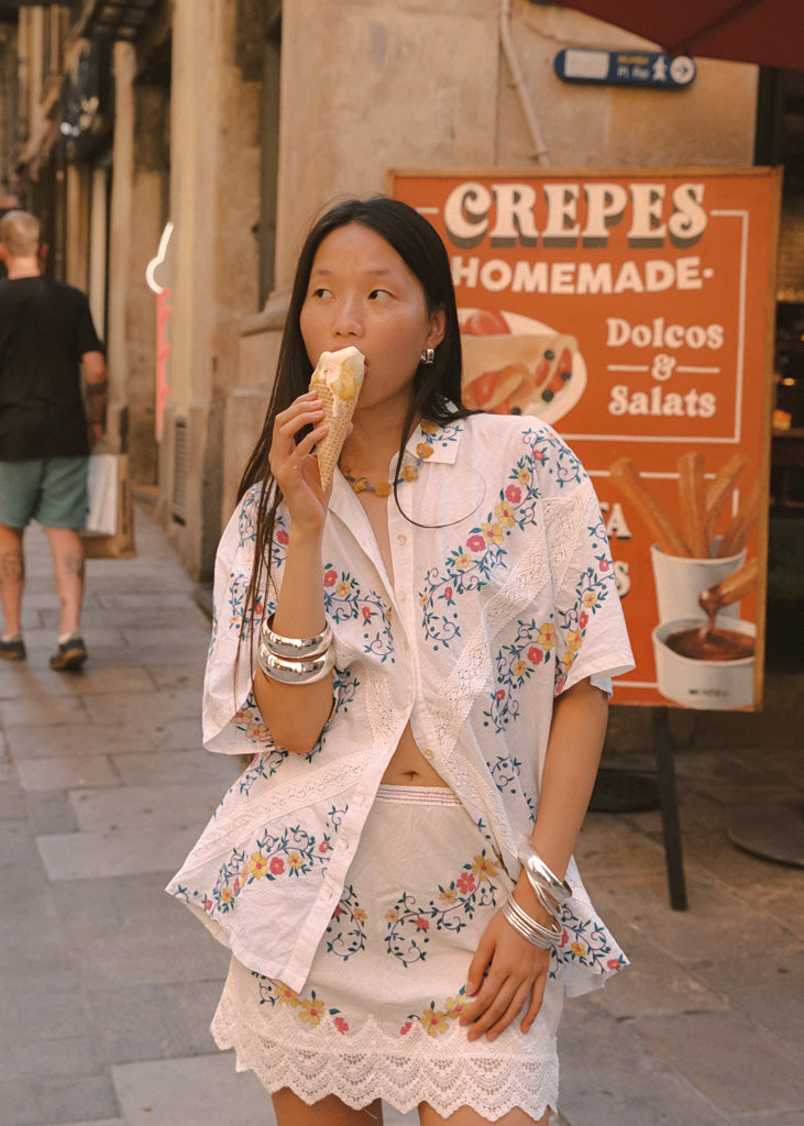 A woman stands on a city street wearing a white embroidered short-sleeve shirt and matching skirt, eating a pastry. A colorful crepe shop sign is visible behind her.