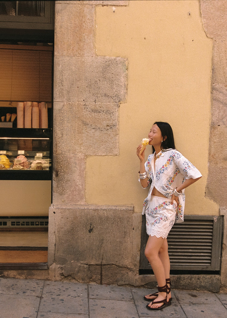 A woman stands on a sidewalk beside a textured beige and gray stone wall, wearing a white floral two-piece set with a cropped jacket and matching skirt, holding a pastry near her mouth.