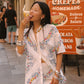 A woman stands on a city street wearing a white embroidered short-sleeve shirt and matching shorts with lace trim. She is eating a pastry in front of a street sign advertising homemade crepes.