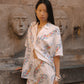 A woman stands outdoors against a stone wall with sculpted faces, wearing a white embroidered short-sleeve shirt and matching mini skirt. The outfit has colorful floral embroidery in pink, blue, and orange tones.