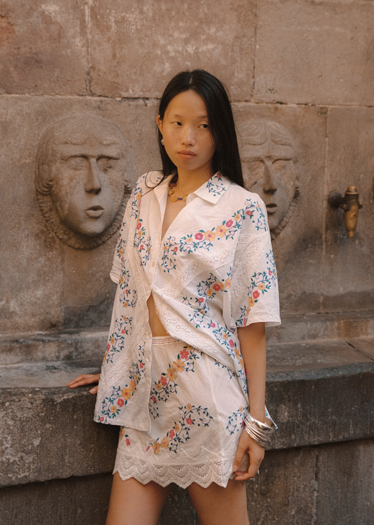 A woman stands outdoors against a stone wall with sculpted faces, wearing a white embroidered short-sleeve shirt and matching mini skirt. The outfit has colorful floral embroidery in pink, blue, and orange tones.
