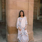 A woman stands against a large tan stone column wearing a loose white caftan dress with colorful floral embroidery and gold trim, along with brown sandals.