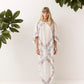 A woman stands against a white wall wearing a long white kaftan dress with colorful floral embroidery. Green houseplants are visible in the corners of the frame.