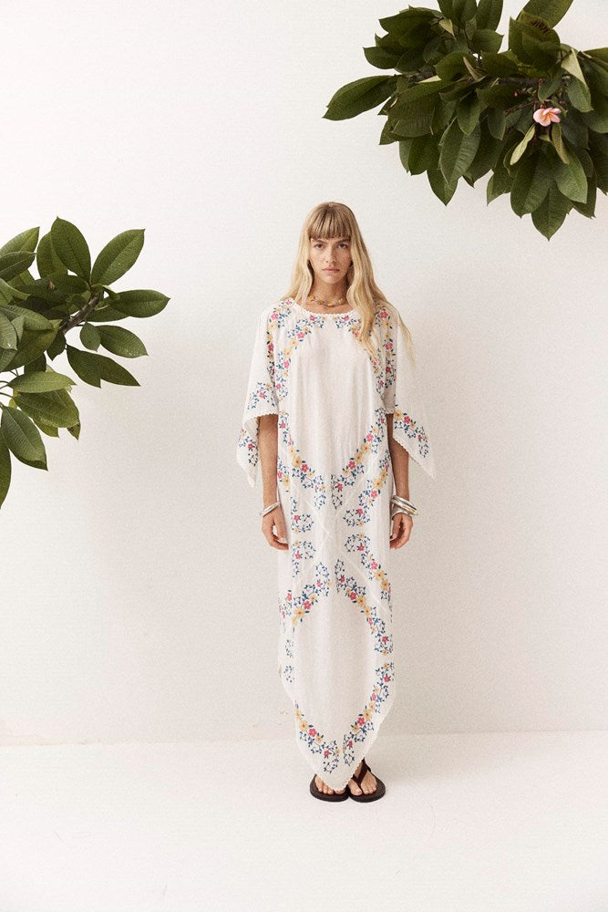 A woman stands against a white wall wearing a long white kaftan dress with colorful floral embroidery. Green houseplants are visible in the corners of the frame.
