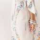 A close-up of a person wearing a flowing white maxi dress with a colorful floral print and lace trim, accessorized with stacked silver bangles. The brand name 'KING A SHILLA' is printed vertically on the skirt.