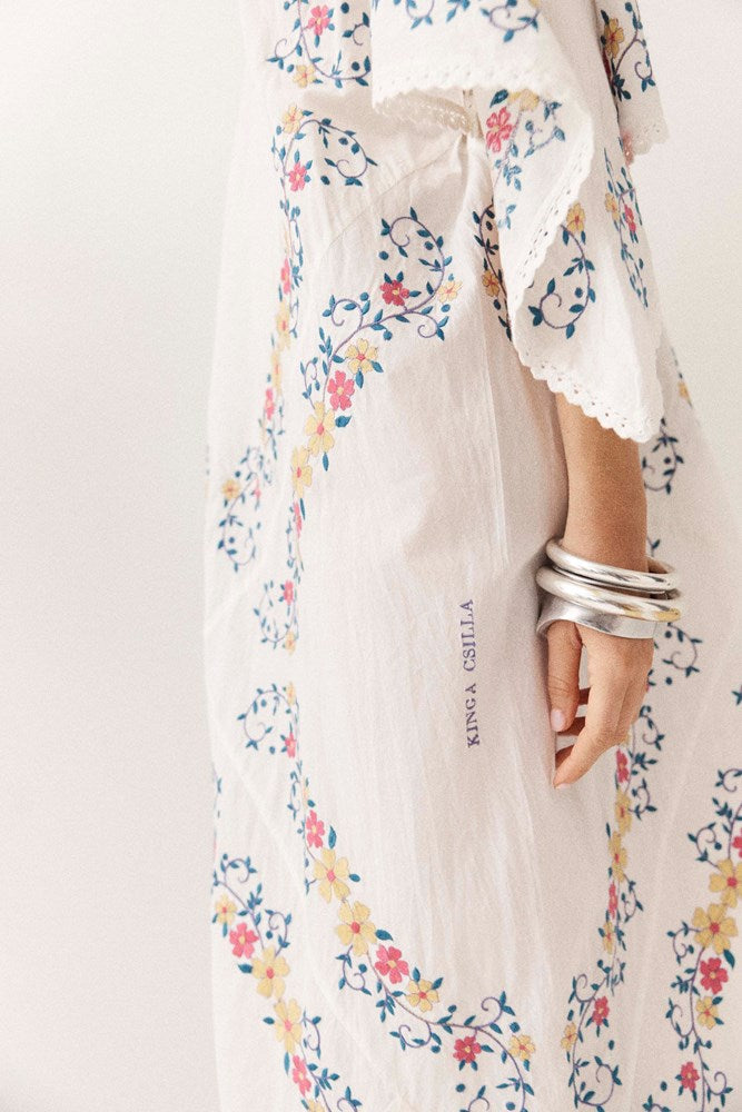 A close-up of a person wearing a flowing white maxi dress with a colorful floral print and lace trim, accessorized with stacked silver bangles. The brand name 'KING A SHILLA' is printed vertically on the skirt.