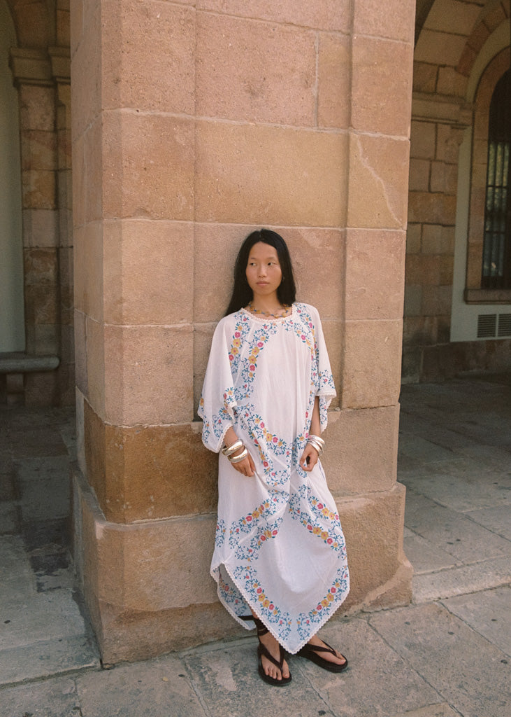 A woman stands against a large tan stone column wearing a loose white caftan dress with colorful floral embroidery and gold trim, along with brown sandals.
