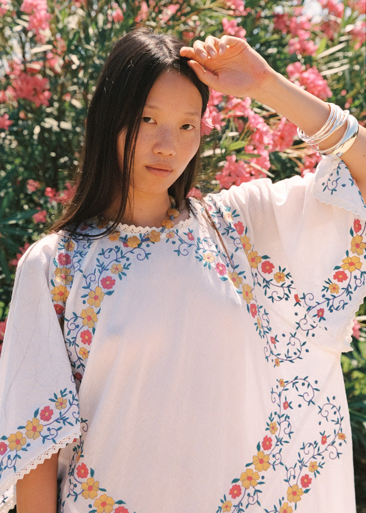 A woman wearing a white bohemian-style tunic dress with colorful floral embroidery, posing outdoors in front of a background of pink flowers.