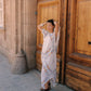A woman wearing a white high-low dress with colorful floral patterns stands in front of a large carved wooden door, posing with her hands behind her head.