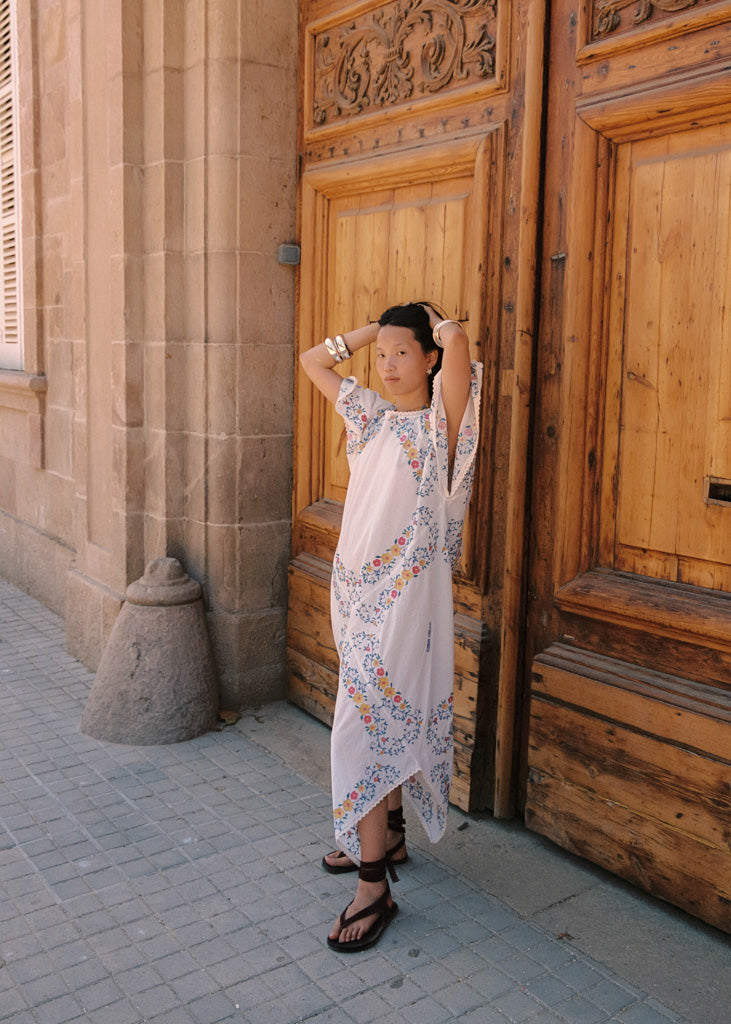A woman wearing a white high-low dress with colorful floral patterns stands in front of a large carved wooden door, posing with her hands behind her head.