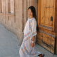 A woman wearing a white bohemian maxi dress with colorful floral embroidery walks along a cobblestone street next to a stone wall and a rustic wooden door.