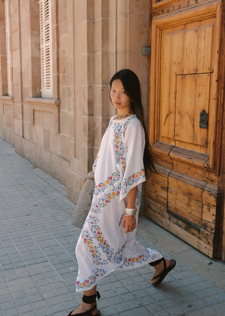 A woman wearing a white bohemian maxi dress with colorful floral embroidery walks along a cobblestone street next to a stone wall and a rustic wooden door.