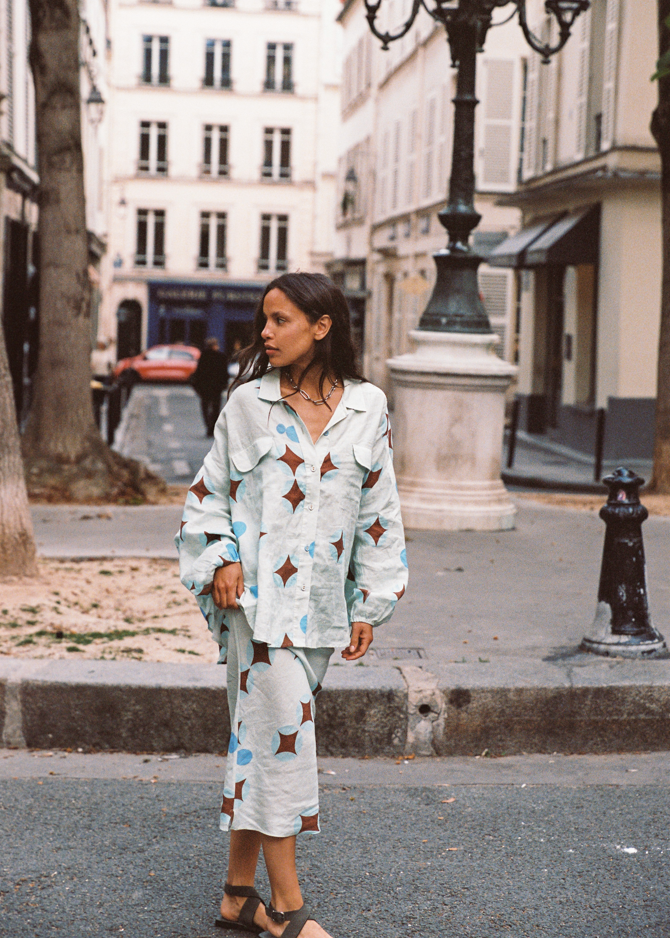 Woman in a light blue celestial patterned shirt and skirt set standing among potted plants and flowers.