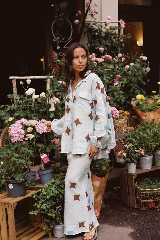 Woman in a light blue celestial patterned shirt and skirt set standing among potted plants and flowers.