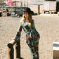 Woman in a brown and blue patterned kimono and flare set in a cowboy hat leaning against a wooden post outdoors.