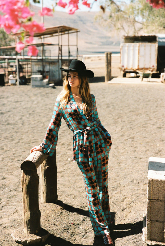 Woman in a brown and blue patterned kimono and flare set in a cowboy hat leaning against a wooden post outdoors.