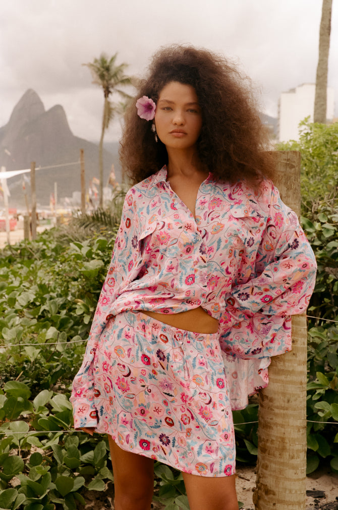 Woman in a pink floral shirt and skirt set standing outdoors with mountains and palm trees in the background