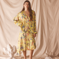 Woman wearing a yellow floral set standing against a beige curtain with decorative elements on the floor.