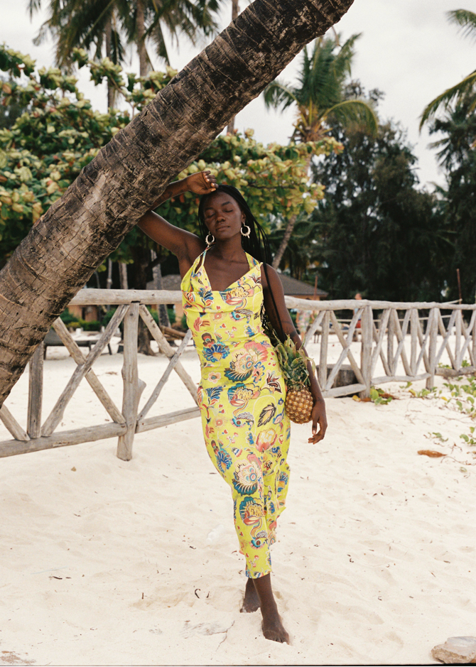 Woman in a yellow floral dress standing under a palm tree on a sandy beach.