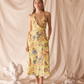 Woman wearing a yellow floral dress standing against a beige curtain with decorative elements on the floor.