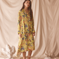 Woman wearing a yellow floral set standing against a beige curtain with decorative elements on the floor.