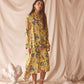 Woman wearing a yellow floral set standing against a beige curtain with decorative elements on the floor.