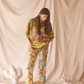 Person wearing a lime green patterned shirt and colorful pants standing against a beige curtain with decorative elements.