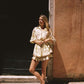 A woman stands outdoors against a warm terracotta wall and dark doorway, wearing a cream wrap tee with ornate gold embroidery and dark brown flat shoes.