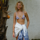 Woman in a floral pink bikini top and blue and white sarong standing against a light-colored wall with a plant in the foreground.