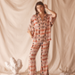 Woman wearing a pink patterned shirt and flare pants standing against a beige curtain with decorative elements.