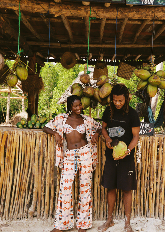 Two people standing in front of a rustic wooden structure with coconuts and tropical plants. Women is wearing pink patterned set and bikini top. 