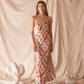 Woman in a pink floral dress standing against a beige curtain with decorative elements on the floor.