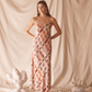 Woman in a pink floral dress standing against a beige curtain with decorative elements on the floor.