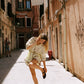 A woman in a light beige patterned skirt and skirt and black flat shoes poses in a sunlit, narrow European alleyway with warm-toned buildings.