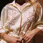 A woman wearing a cream-colored cotton shirt with brown embroidered sun motifs and a rope tie belt, shown in a warm, sunlit setting.