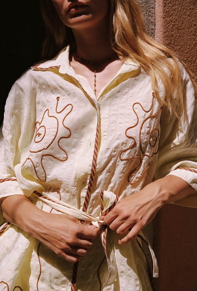 A woman wearing a cream-colored cotton shirt with brown embroidered sun motifs and a rope tie belt, shown in a warm, sunlit setting.