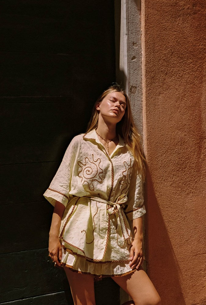 A woman wearing a cream-colored embroidered short-sleeve tunic dress with a tie waist, leaning against a textured terracotta wall next to a dark doorway.