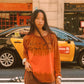 A woman stands on a city street wearing an orange long-sleeve t-shirt with 'Kinga Csilla' printed across the chest and brown shorts with orange trim. A yellow and black taxi is blurred in the background.