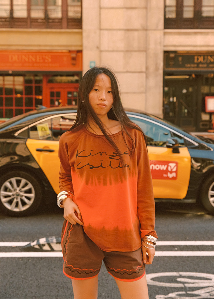 A woman stands on a city street wearing an orange long-sleeve t-shirt with 'Kinga Csilla' printed across the chest and brown shorts with orange trim. A yellow and black taxi is blurred in the background.