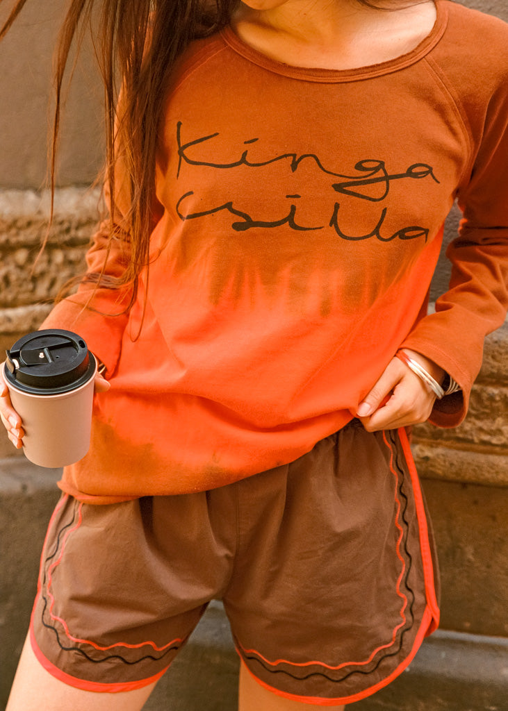 A person wearing a burnt orange long-sleeve t-shirt with 'Kinga Csilla' printed in black script, paired with brown shorts featuring orange and black wavy trim. The person is holding a coffee cup, standing outdoors against a stone wall.