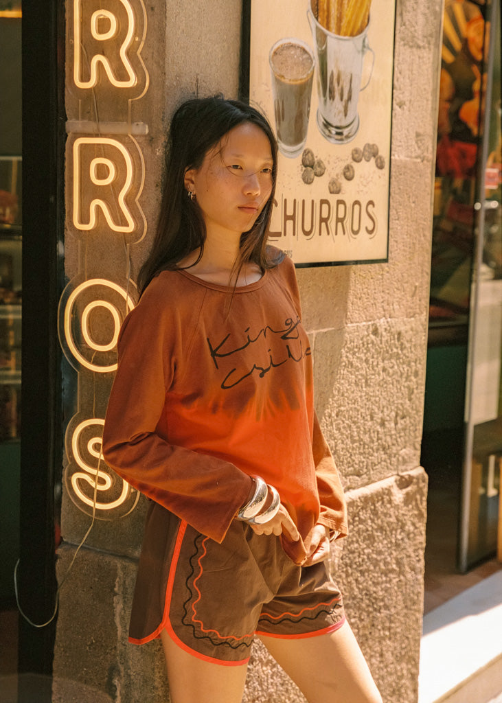 A woman stands outdoors against a stone wall next to a neon sign, wearing a rust-orange long-sleeve t-shirt with 'Kinga Csilla' printed in black script and brown shorts with orange trim.