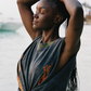 Woman with braided hair wearing a dark gray tank with colorful patterns, standing by water.