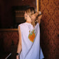 A woman viewed from behind wearing a sleeveless light blue top with a colorful flame graphic on the back, standing in a room with ornate brown patterned wallpaper and a large mirror.