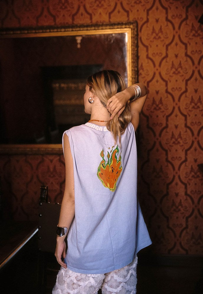 A woman viewed from behind wearing a sleeveless light blue top with a colorful flame graphic on the back, standing in a room with ornate brown patterned wallpaper and a large mirror.