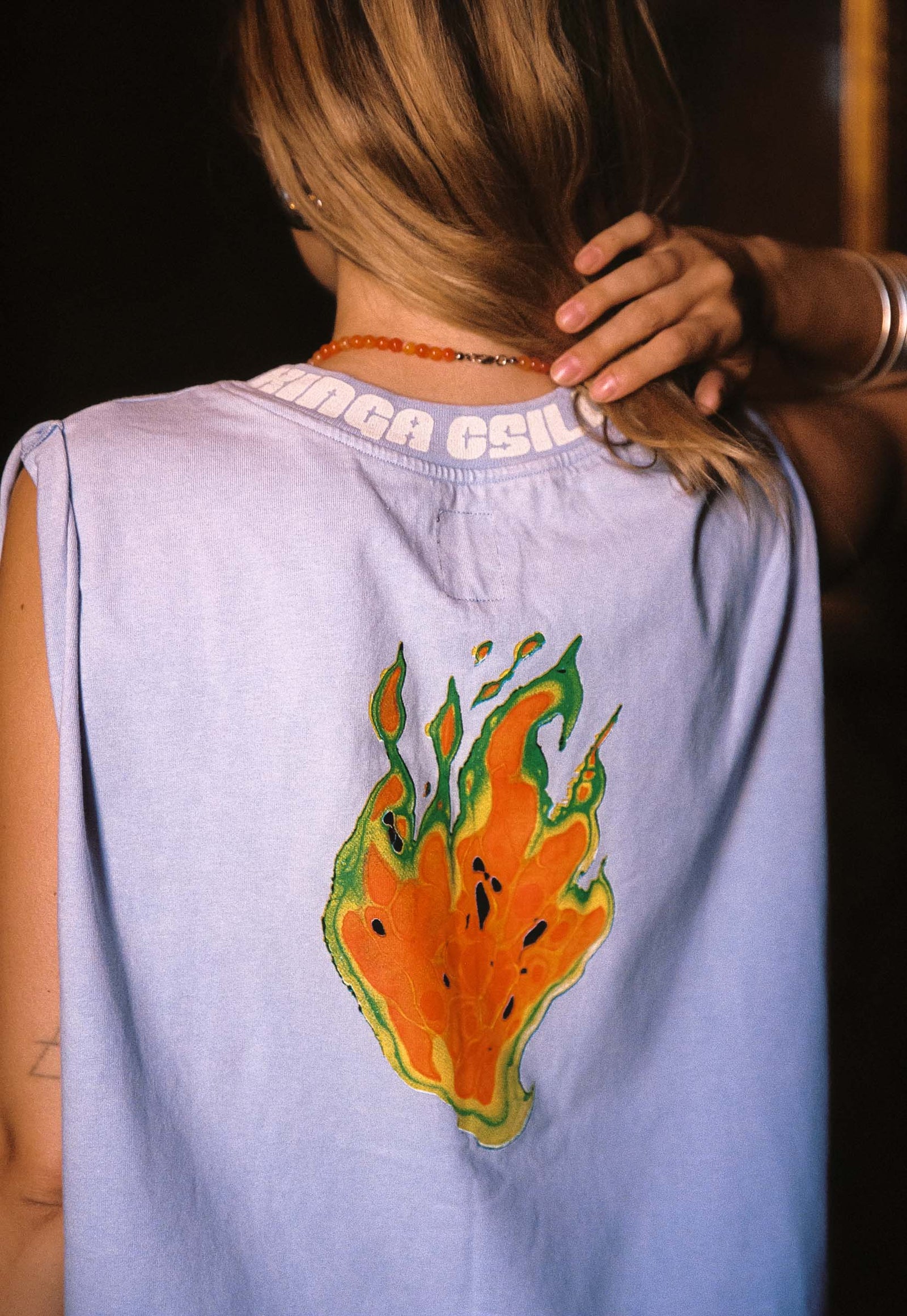 A person shown from the back wearing a sleeveless periwinkle top with a large orange and green flame graphic printed on the back. The brand name 'KINGA CSILLA' is printed in white along the inside neckline.