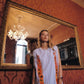 A woman wearing a light blue sleeveless tunic with orange and white patterned side panels stands in front of a large ornate gold-framed mirror in a room with red patterned wallpaper.