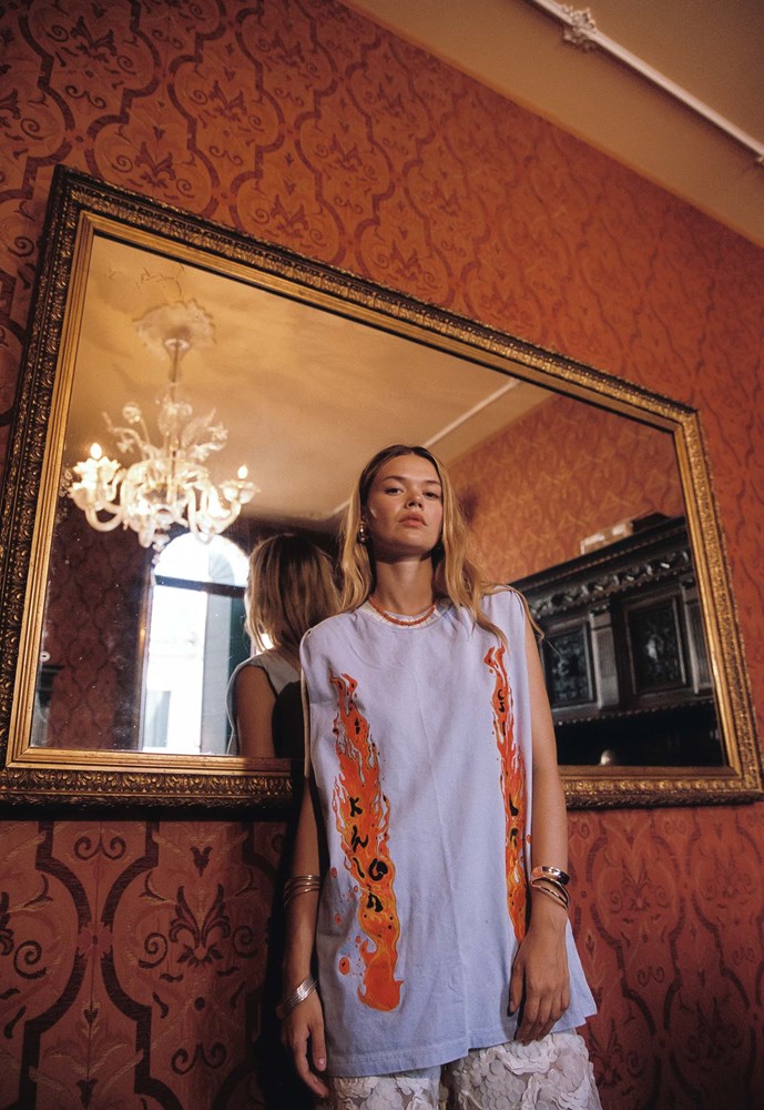 A woman wearing a light blue sleeveless tunic with orange and white patterned side panels stands in front of a large ornate gold-framed mirror in a room with red patterned wallpaper.
