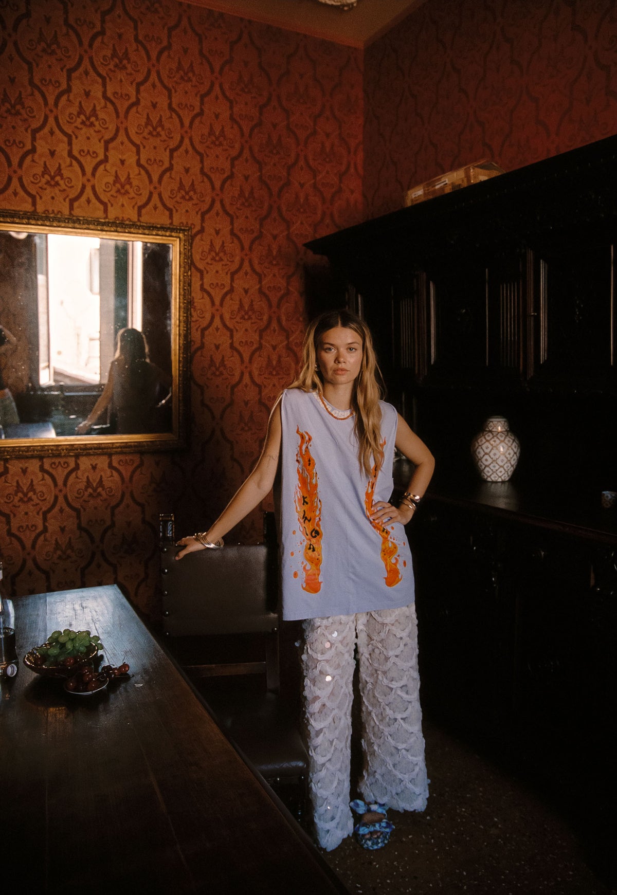 A woman stands in a dimly lit room with ornate red damask wallpaper, wearing a light blue sleeveless top with orange graphic prints and white sequin pants.