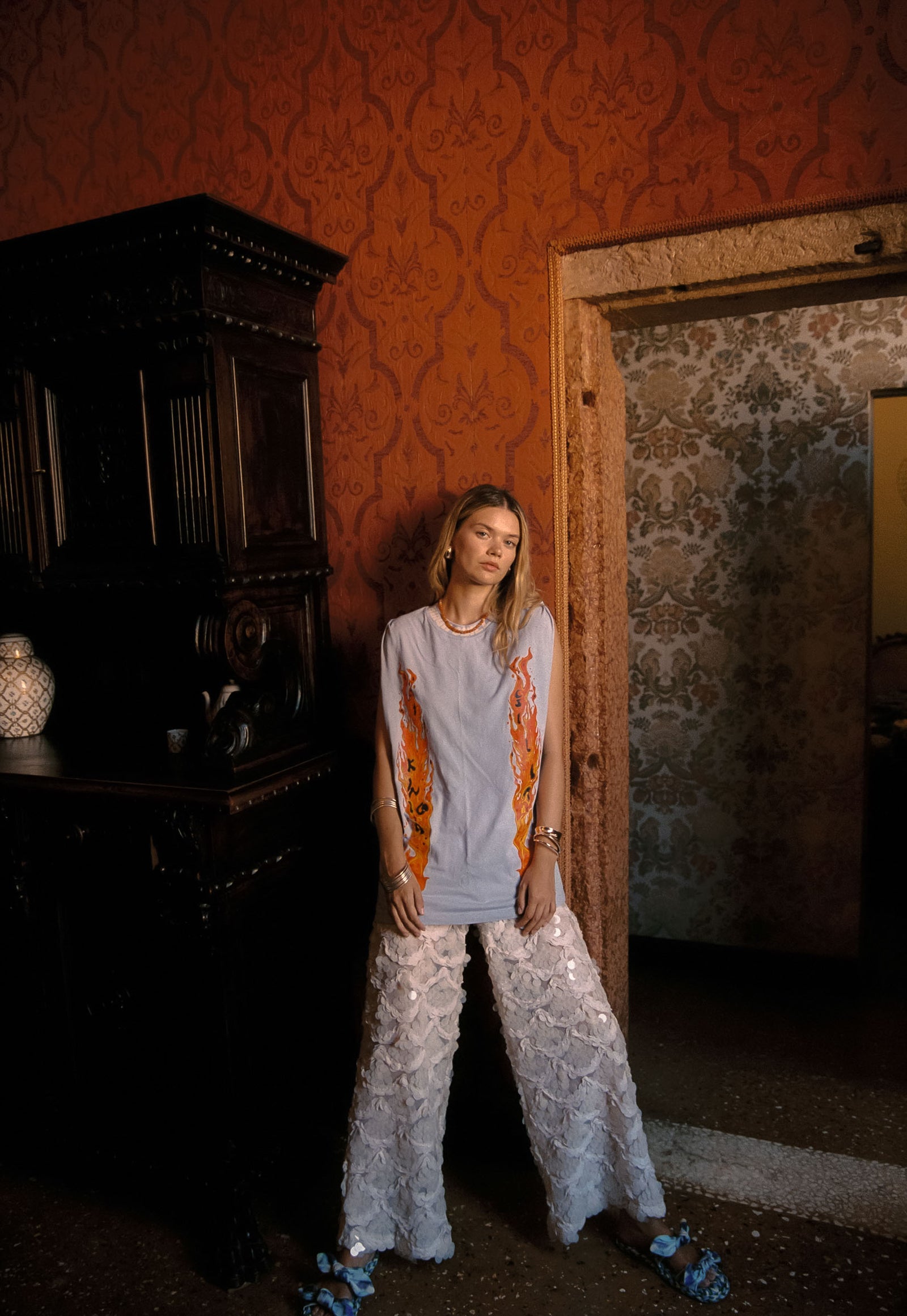 A woman stands in a doorway wearing a periwinkle oversized tank top with orange abstract vertical prints and white sequin wide-leg pants, posed in a dimly lit room with ornate orange wallpaper and dark wood furniture.